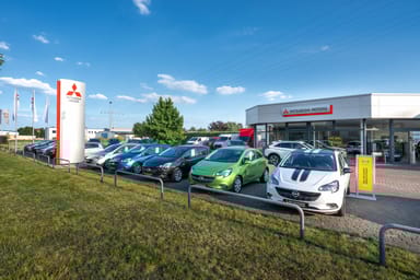 Mitsubishi- und Opel-Fahrzeuge auf dem Gelände des Autohauses Strauch in Rüdersdorf, mit einem Mitsubishi Motors-Schild und dem Autohaus im Hintergrund